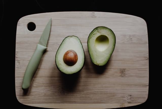 haas avocado cut in half beside a knife sitting on a bamboo cutting board